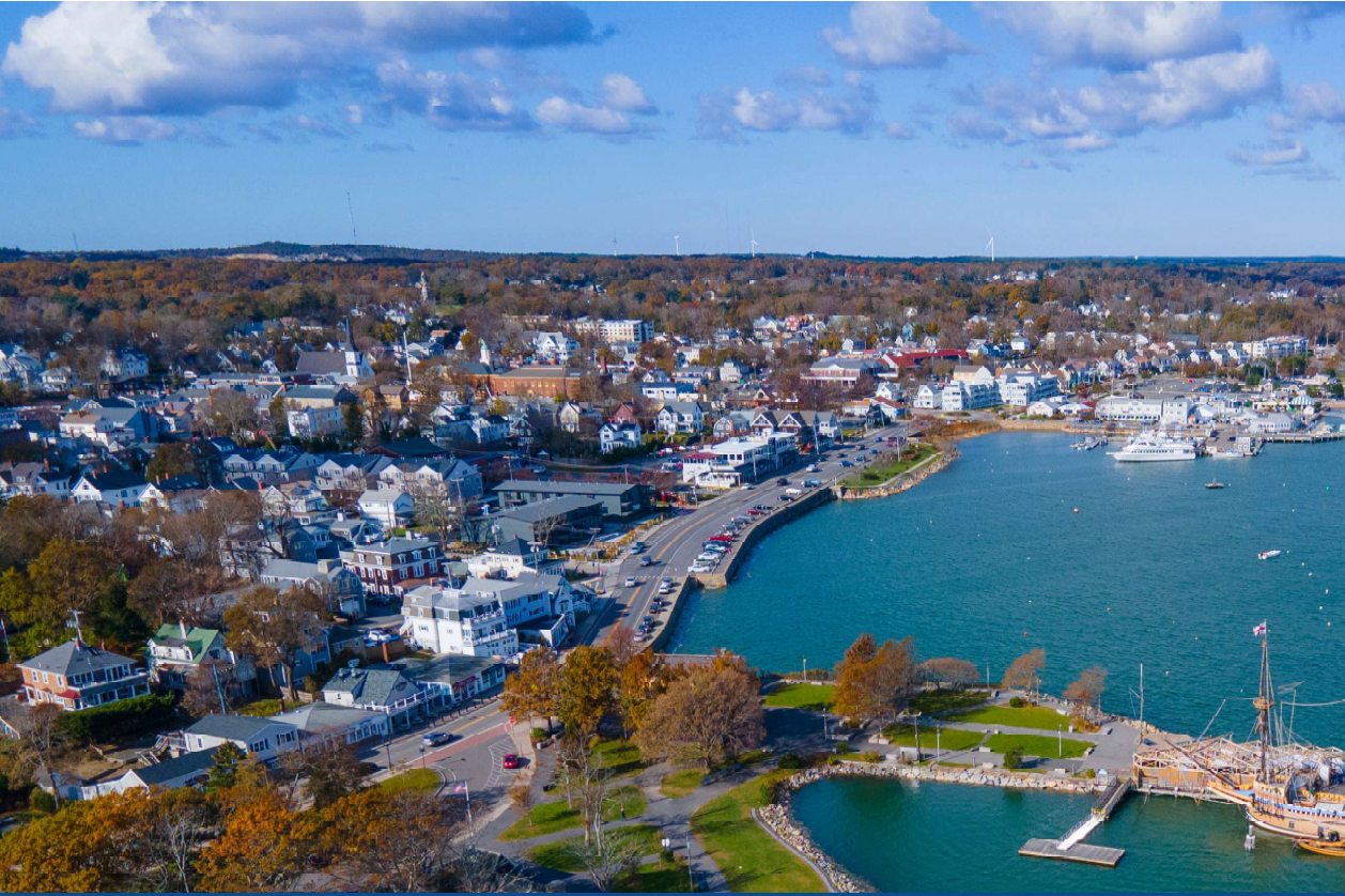 Aerial view of a picturesque New England coastal town in autumn, featuring a curved harbor with turquoise blue water surrounded by residential neighborhoods with fall foliage. The scene shows white and colorful houses dotting tree-lined streets